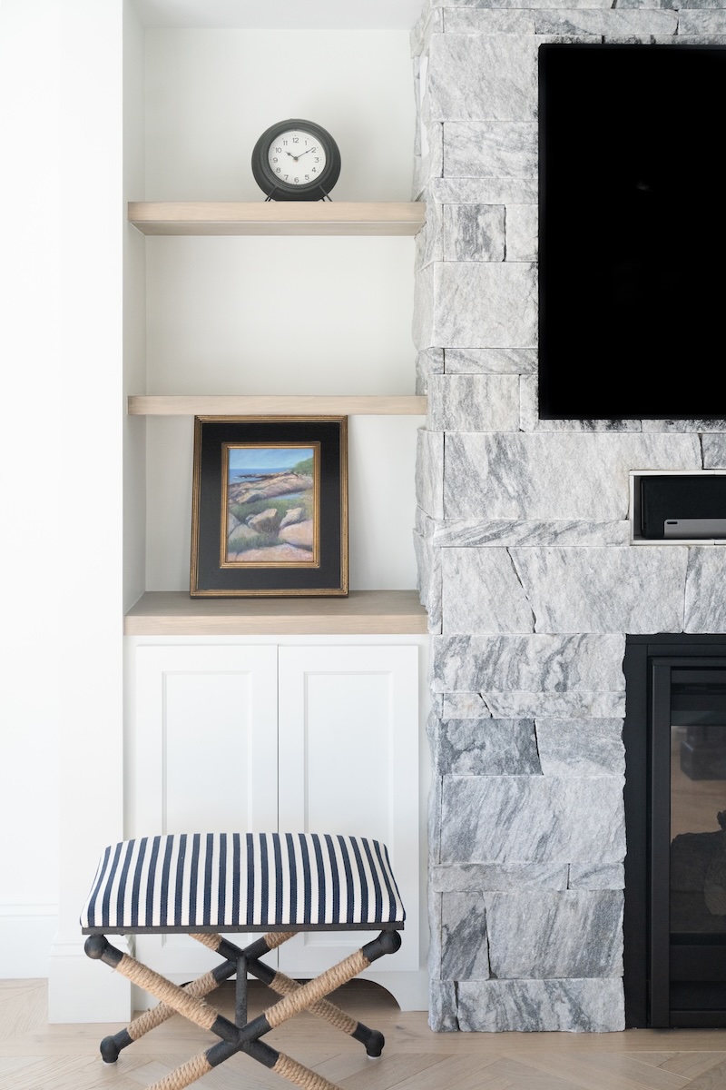 Georgia Marble Pearl Grey™ Berkshire thin veneer fireplace with flanking built-in shelving — coastal interior living room with herringbone oak floor and striped accent stool