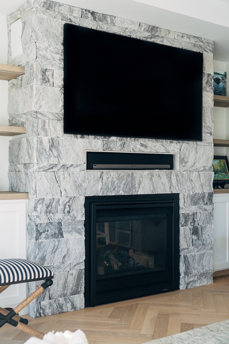 Full view of Berkshire™ Georgia Marble Pearl Grey thin veneer fireplace wall with mounted TV — split-face marble veneer surround, herringbone hardwood floor, coastal interior design